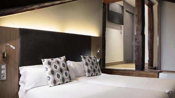 Un lit bien fait, avec du linge de lit blanc et des coussins à motifs, une tête de lit en bois et un grand miroir qui reflète l’intérieur de la pièce.