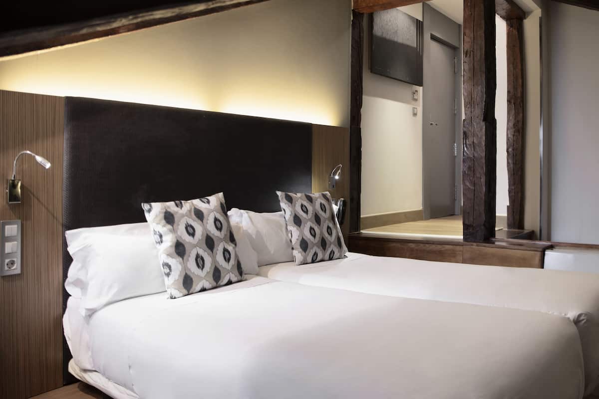 Una cama bien hecha con sábanas blancas y almohadas estampadas, un cabecero de madera y un espejo grande que refleja el interior de la habitación.