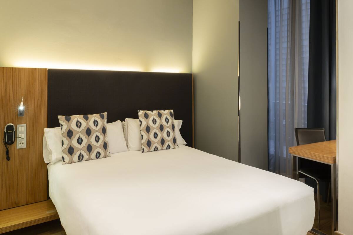 Una habitación de hotel con una cama, una mesita de noche de madera, un escritorio y una silla.