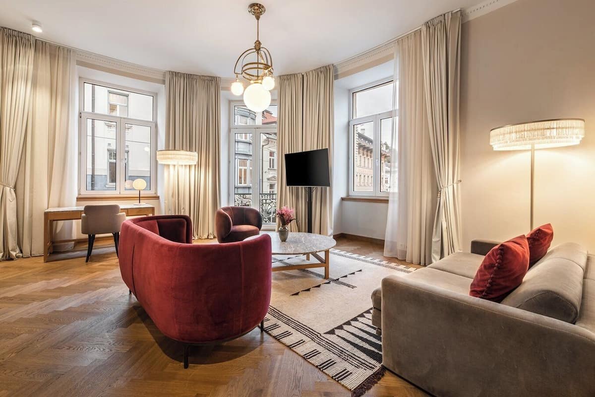 A modern living room with a red armchair, a grey sofa, and a TV. There are large windows with curtains, a chandelier, and a floor lamp.