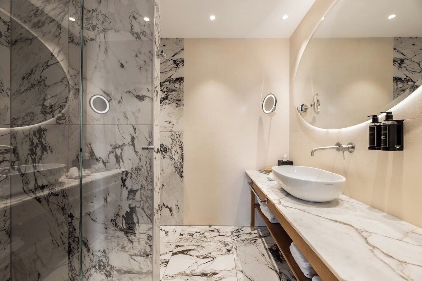 A modern bathroom with a glass shower enclosure, a marble-tiled floor, and a large oval sink with a mirror above it.