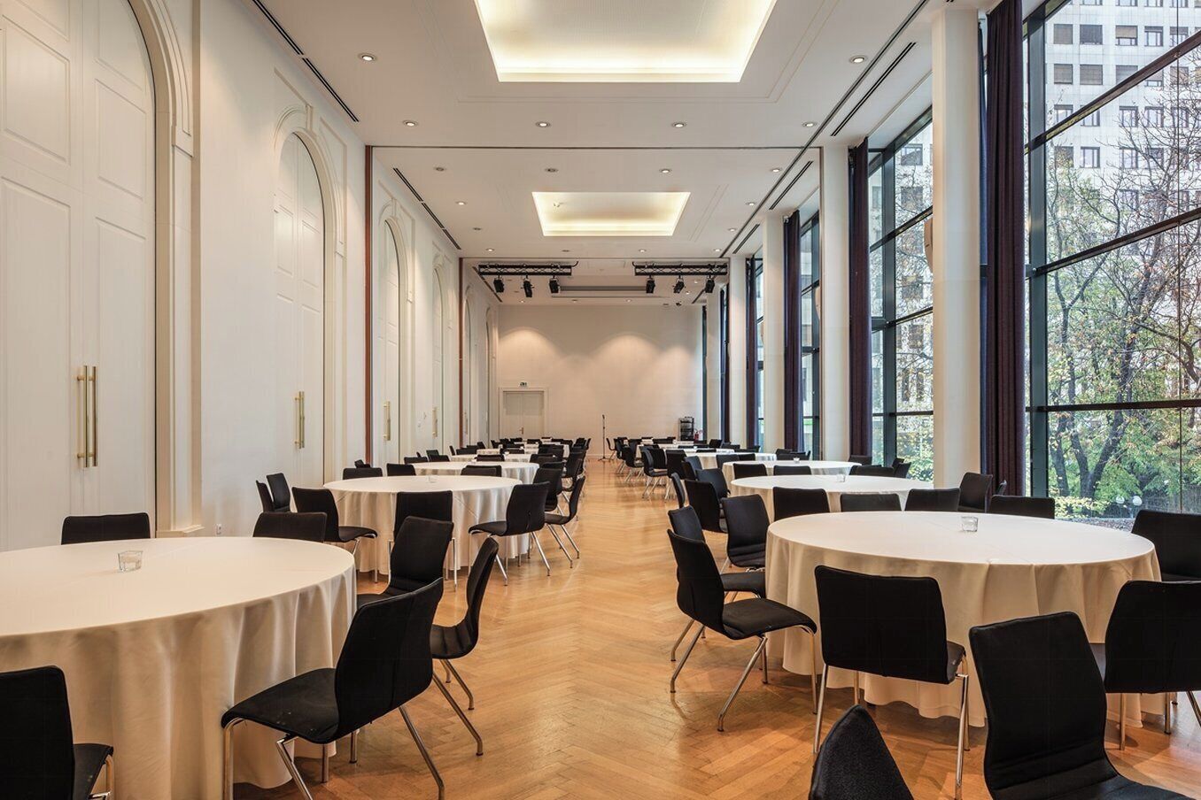 Un salón de conferencias con varias mesas redondas y sillas negras, ventanas grandes y suelo de madera.