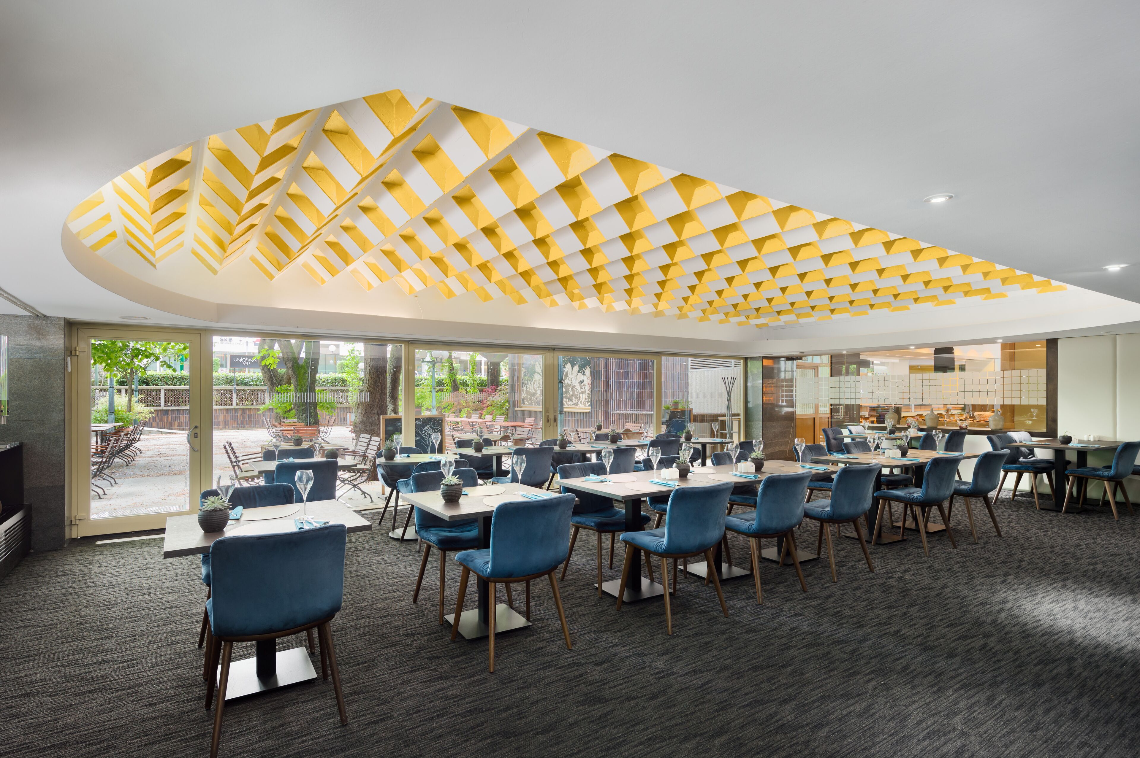 A modern dining area with tables and chairs, a patterned ceiling, and large windows offering views of the outdoors.
