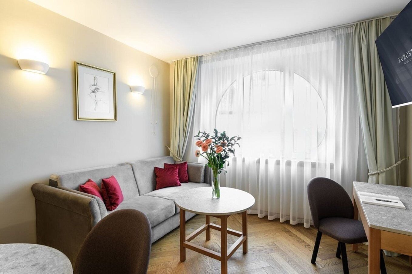 A modern living room with a grey sofa, red cushions, a round wooden table, and a vase of flowers.