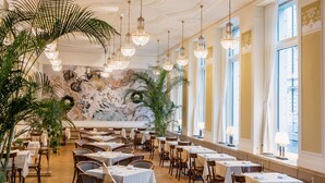 Uma sala de jantar ampla, com várias mesas e cadeiras, lustres elegantes e grandes janelas que permitem entrada de luz natural.
