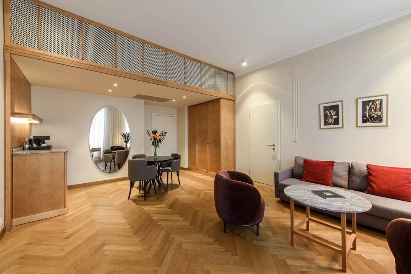 A modern living room with a herringbone wooden floor, a round dining table with chairs, a sofa with red cushions, and a coffee machine on a wooden counter.
