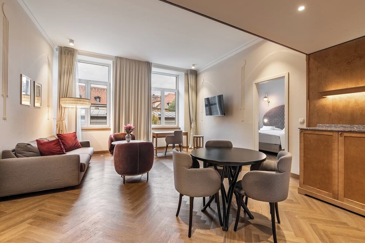 A modern hotel room with a sofa, dining table, chairs, a TV, and a kitchen area.