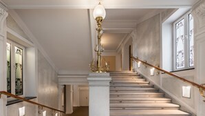 Uma escadaria imponente com corrimão branco, um pilar central e um lustre decorativo.