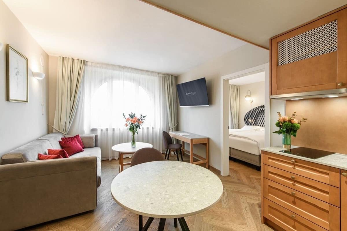 A modern hotel room with a sofa, round dining table, and a kitchenette.