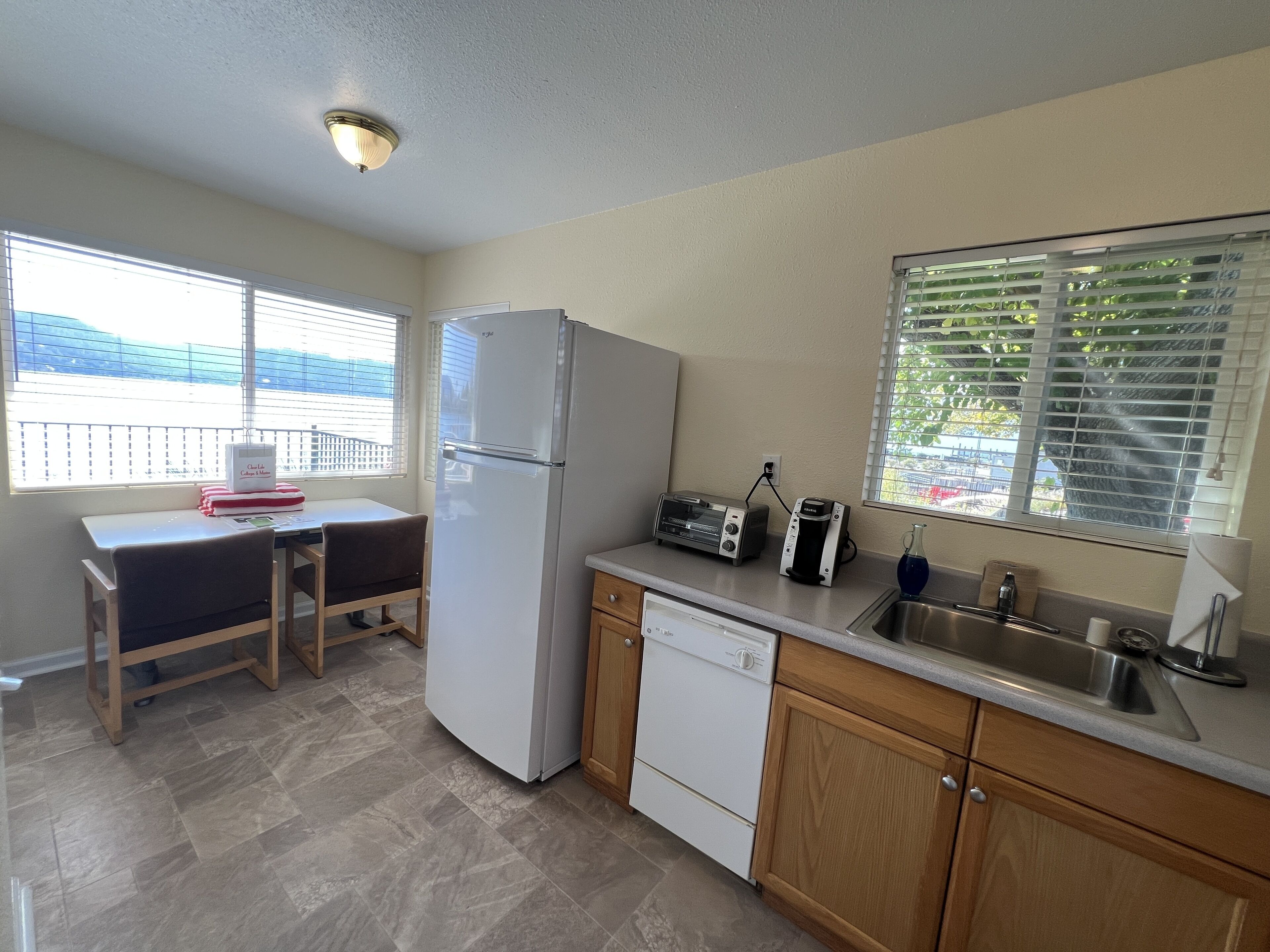 A compact kitchen with a refrigerator, toaster, and dishwasher. There is a window with blinds, a small table, and a chair.