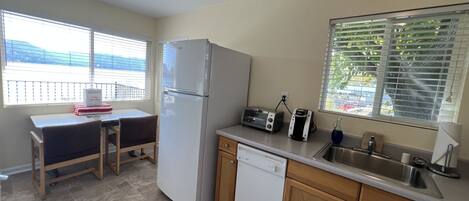 A compact kitchen with a refrigerator, toaster, and dishwasher. There is a window with blinds, a small table, and a chair.