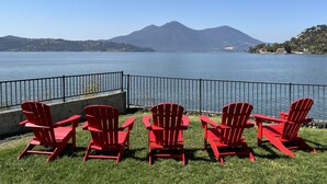 A lakeside view with five red Adirondack chairs facing the water, a metal railing, and a mountain in the background. - Clear Lake Cottages & Marina (Clearlake)