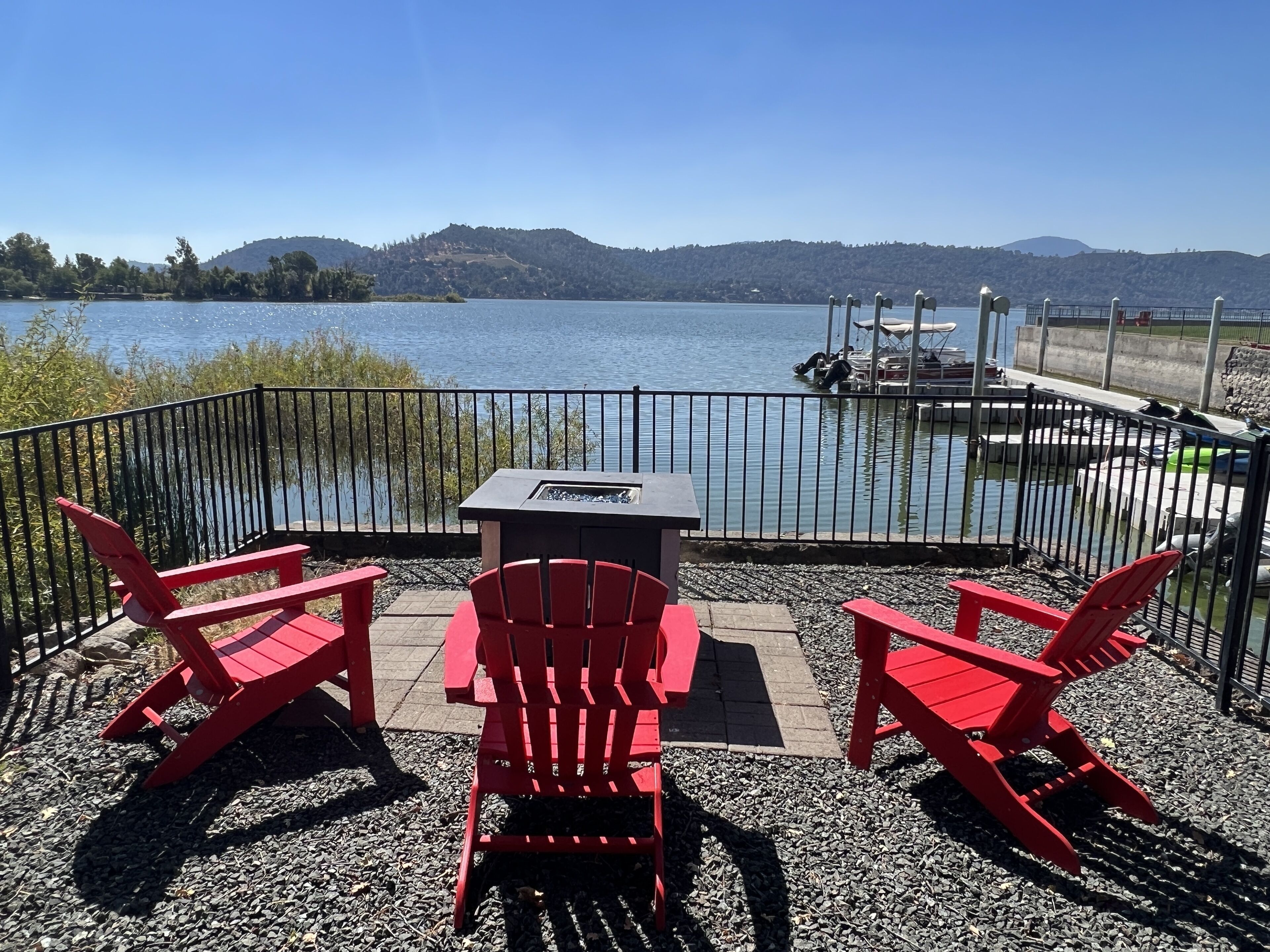 An outdoor seating area with red Adirondack chairs and a table, overlooking a lake with boats and a dock.