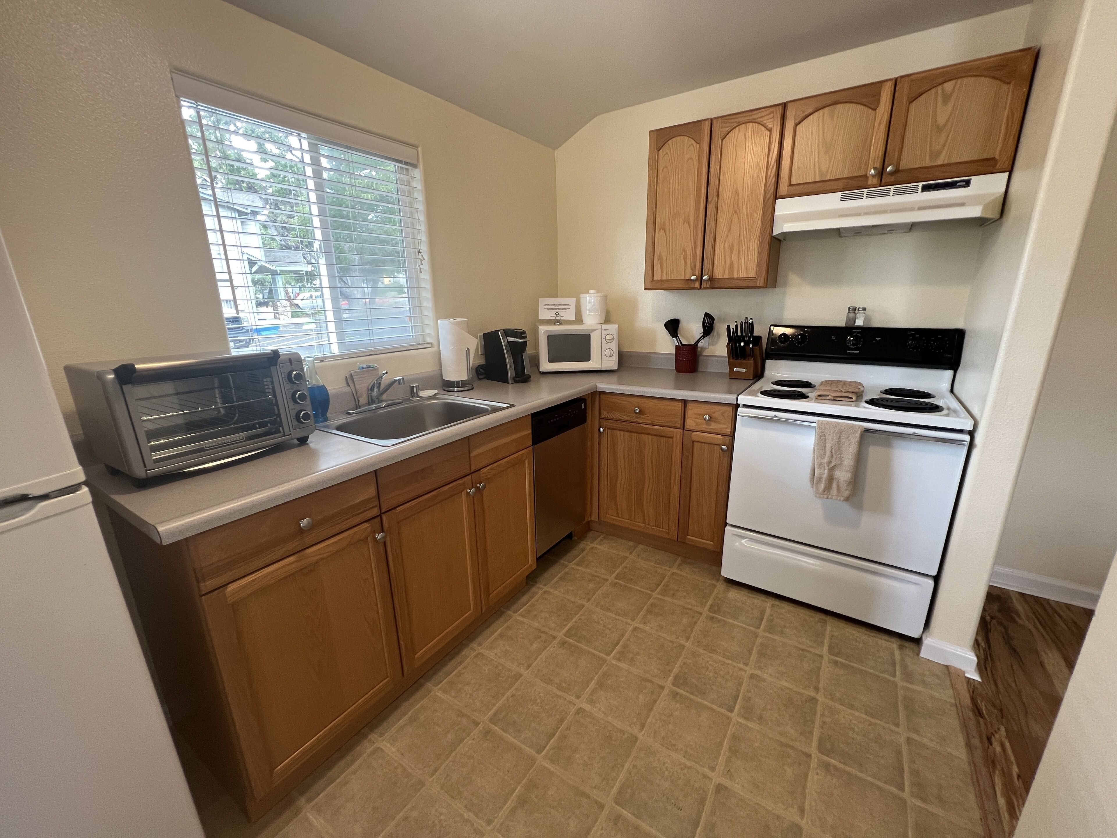 A kitchen with wooden cabinets, a stainless steel sink, a built-in oven, and a window with blinds.
