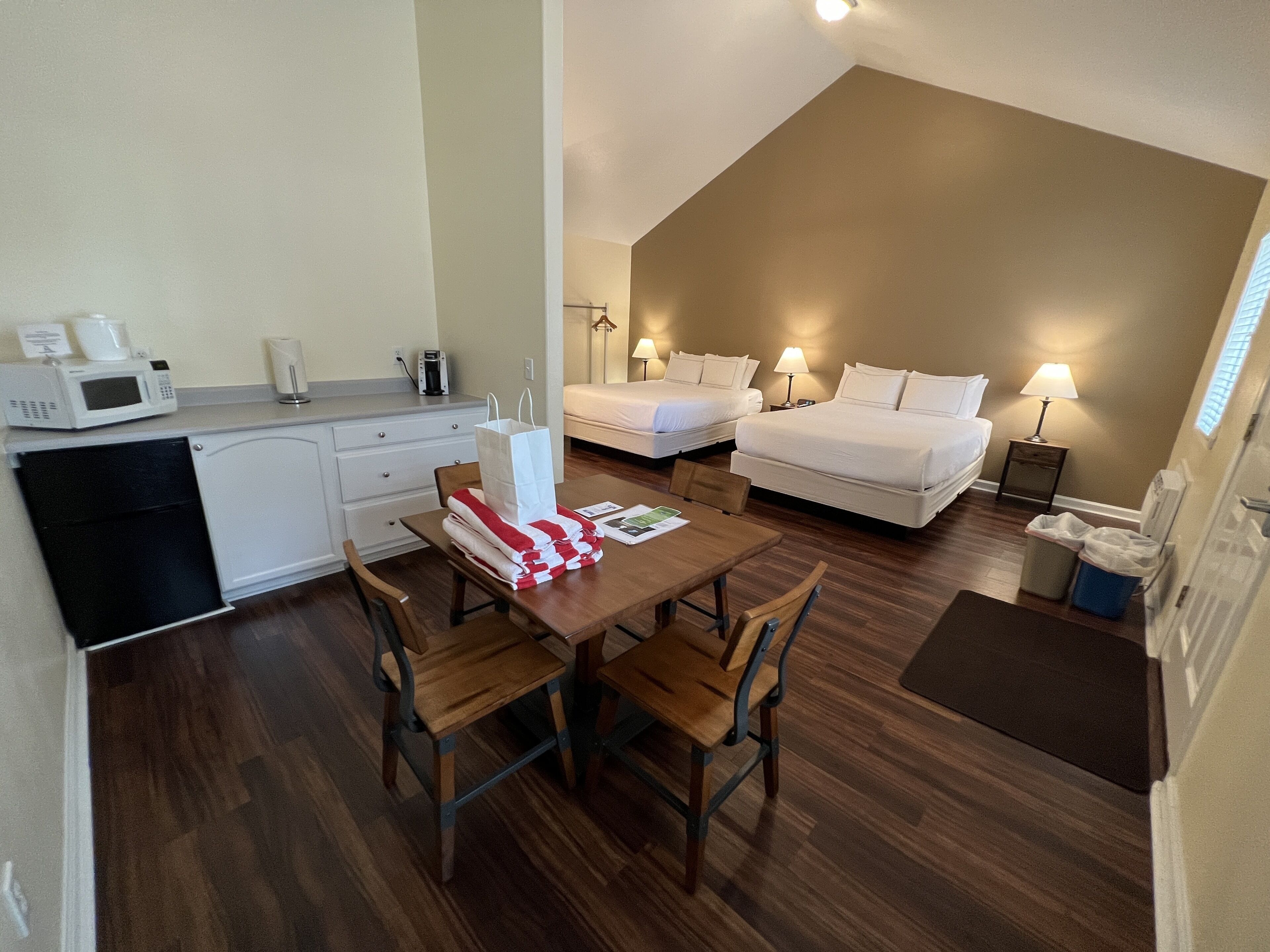 A compact hotel room with a kitchenette, two beds, a dining area, and a small bathroom.