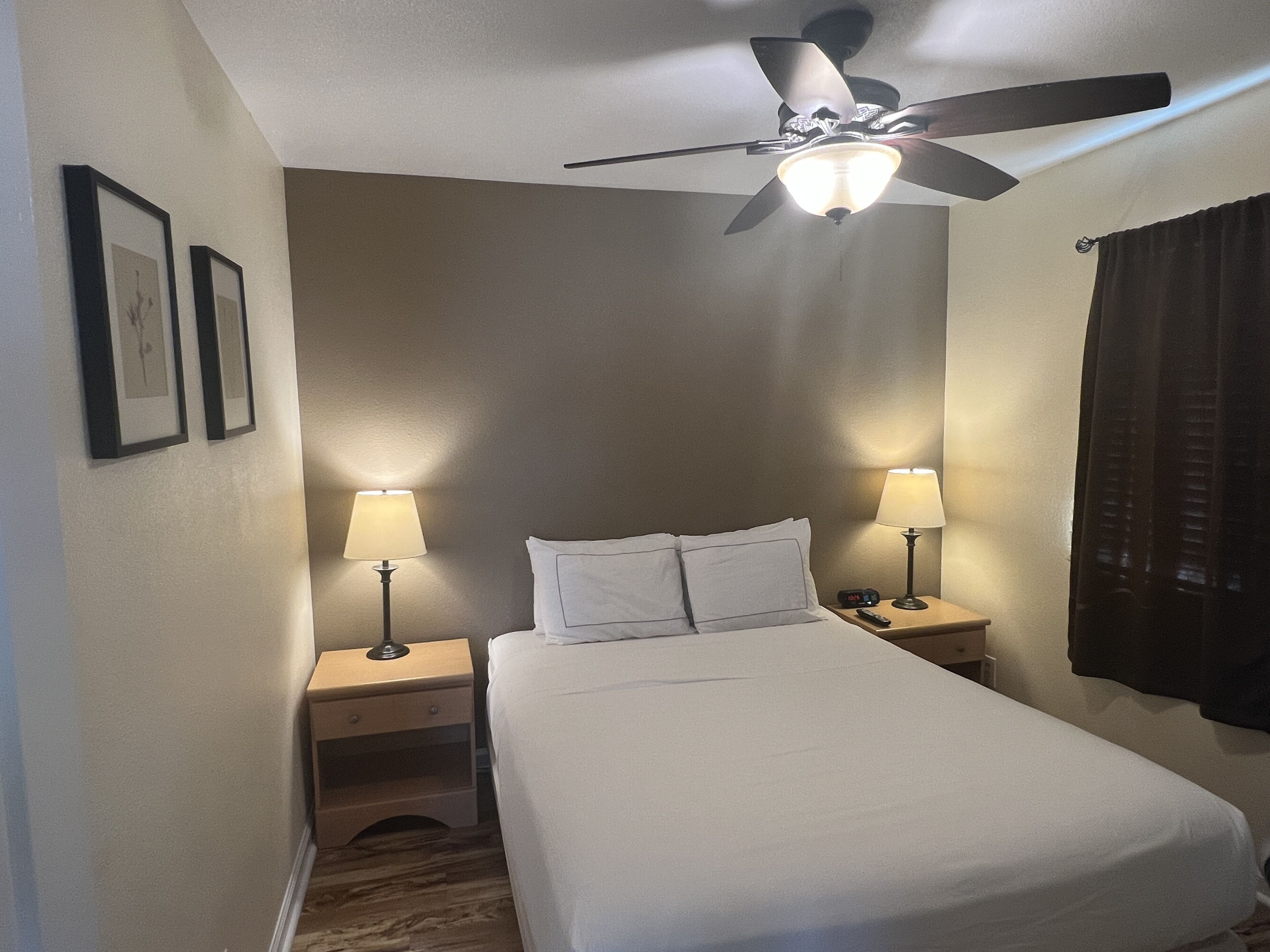 A hotel room with a bed, two bedside tables with lamps, a ceiling fan, and framed pictures on the wall.
