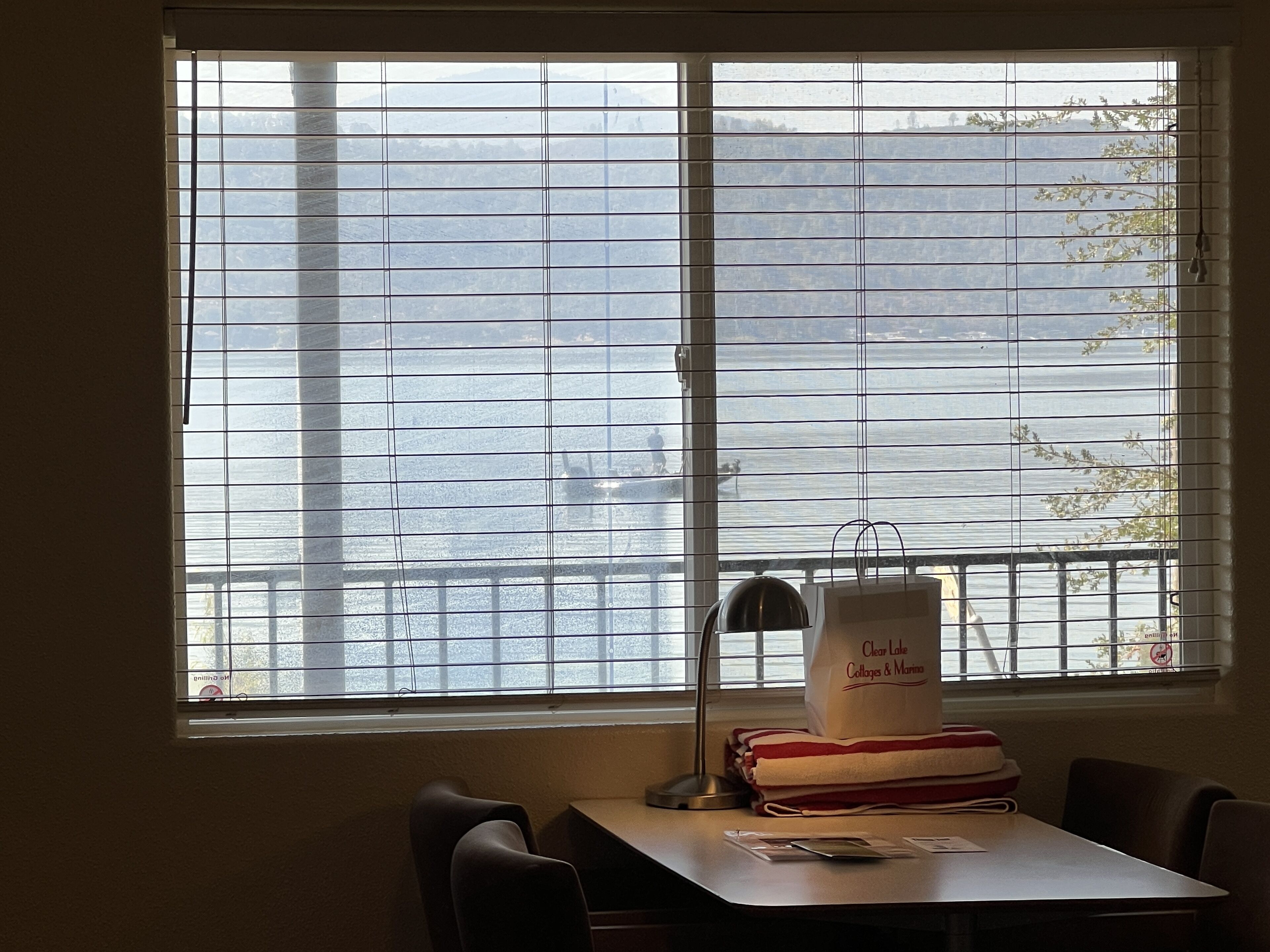 Ein Zimmer mit großem Fenster mit Blick auf ein Gewässer, einem Schreibtisch mit Lampe und einer Tasche mit dem Aufdruck 'Clear Lake Collages & Marina'.