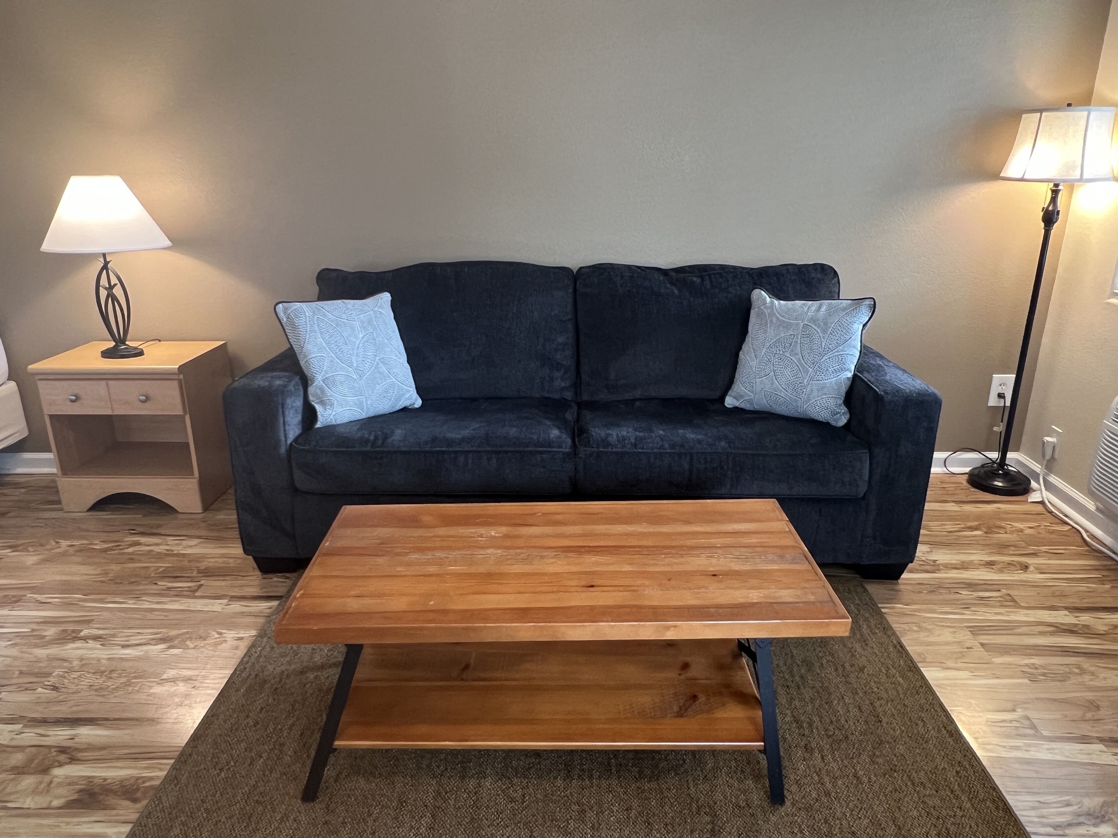 Un salon avec un canapé bleu foncé, deux coussins assortis, une table basse en bois, une table d’appoint avec une lampe et un lampadaire.