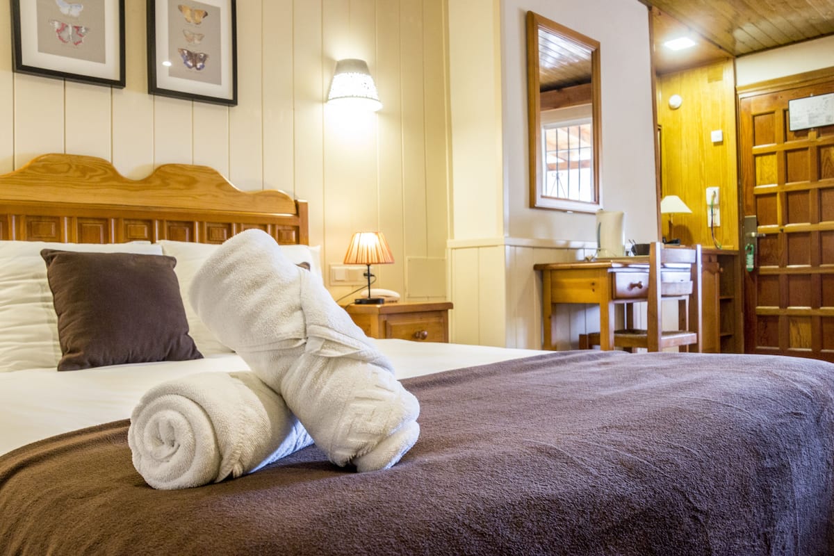 A neatly made bed with a brown blanket and rolled white towels, a wooden headboard, a bedside lamp, and a framed picture on the wall.