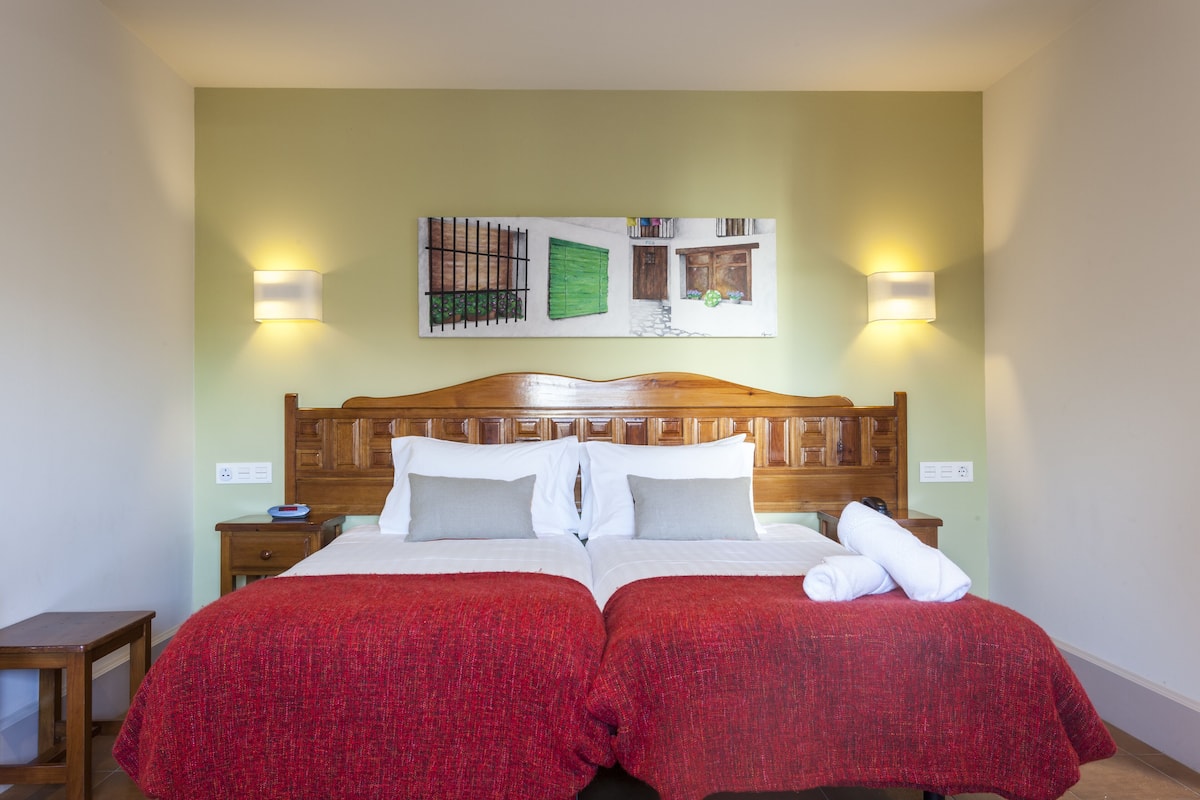 A double bed with red bedding, two bedside tables, a wooden headboard, and a wall-mounted artwork.