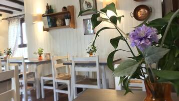 Zona pranzo con sedie e tavoli di legno bianco, un vaso con una pianta e una parete con mensole che ospitano diversi oggetti.
