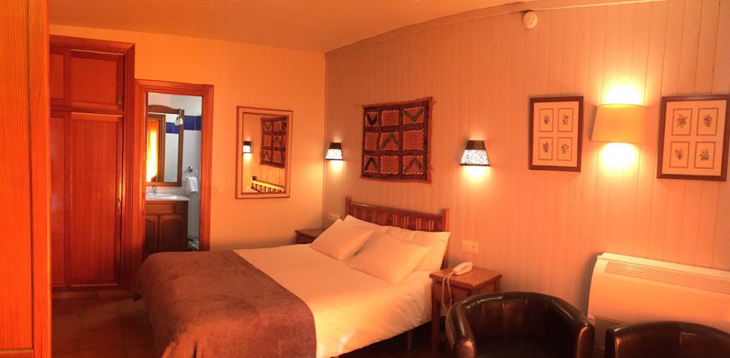 Una habitación de hotel con cama, mesitas de noche, una silla y lámparas fijadas a la pared.
