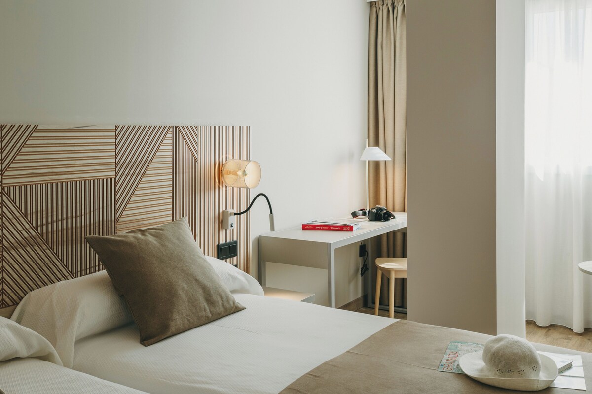 A hotel room with a bed, a desk, a lamp, and a hat on the floor.