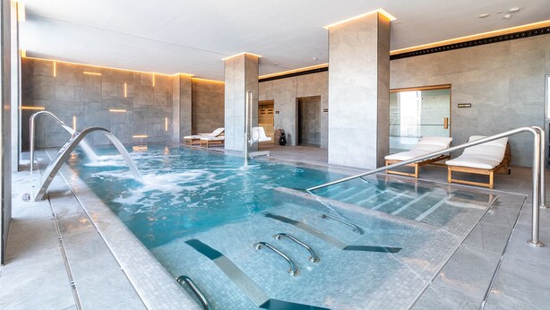 Una piscina interior moderna con agua cristalina, barandillas metálicas y sillas de descanso.