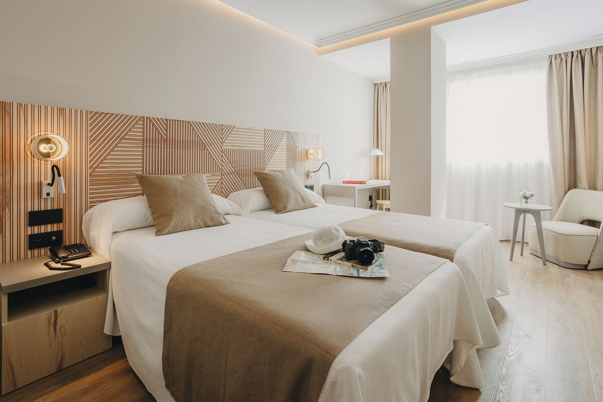 Habitación de hotel con dos camas, un escritorio, una silla, una lámpara y un teléfono en la mesita de noche.