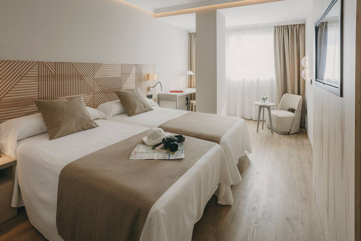 Une chambre d’hôtel avec deux lits, un bureau, une chaise et une grande fenêtre avec des rideaux.