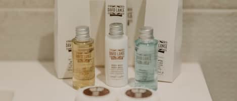 Un baño con encimera blanca, lavabo blanco y espejo. Hay tres botellas de leche corporal David Lance y dos objetos marrones y redondos sobre la encimera.