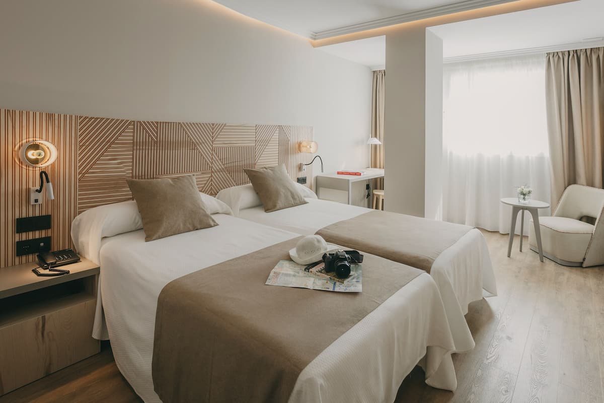 Une chambre d’hôtel avec deux lits, un bureau, une chaise et une grande fenêtre avec des rideaux.