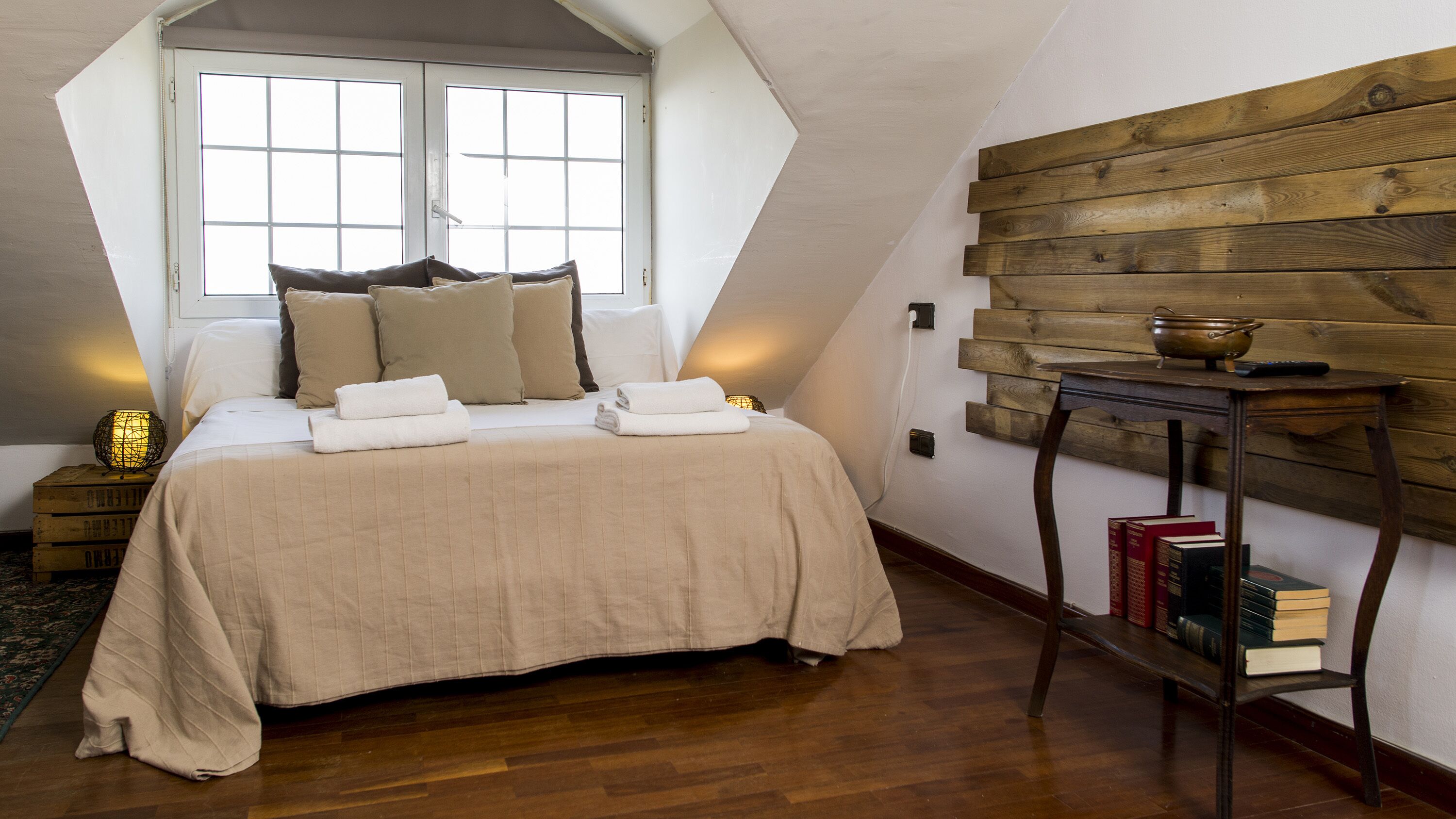 A bedroom with a large bed, a wooden wall panel, and a bedside table with books and a lamp.
