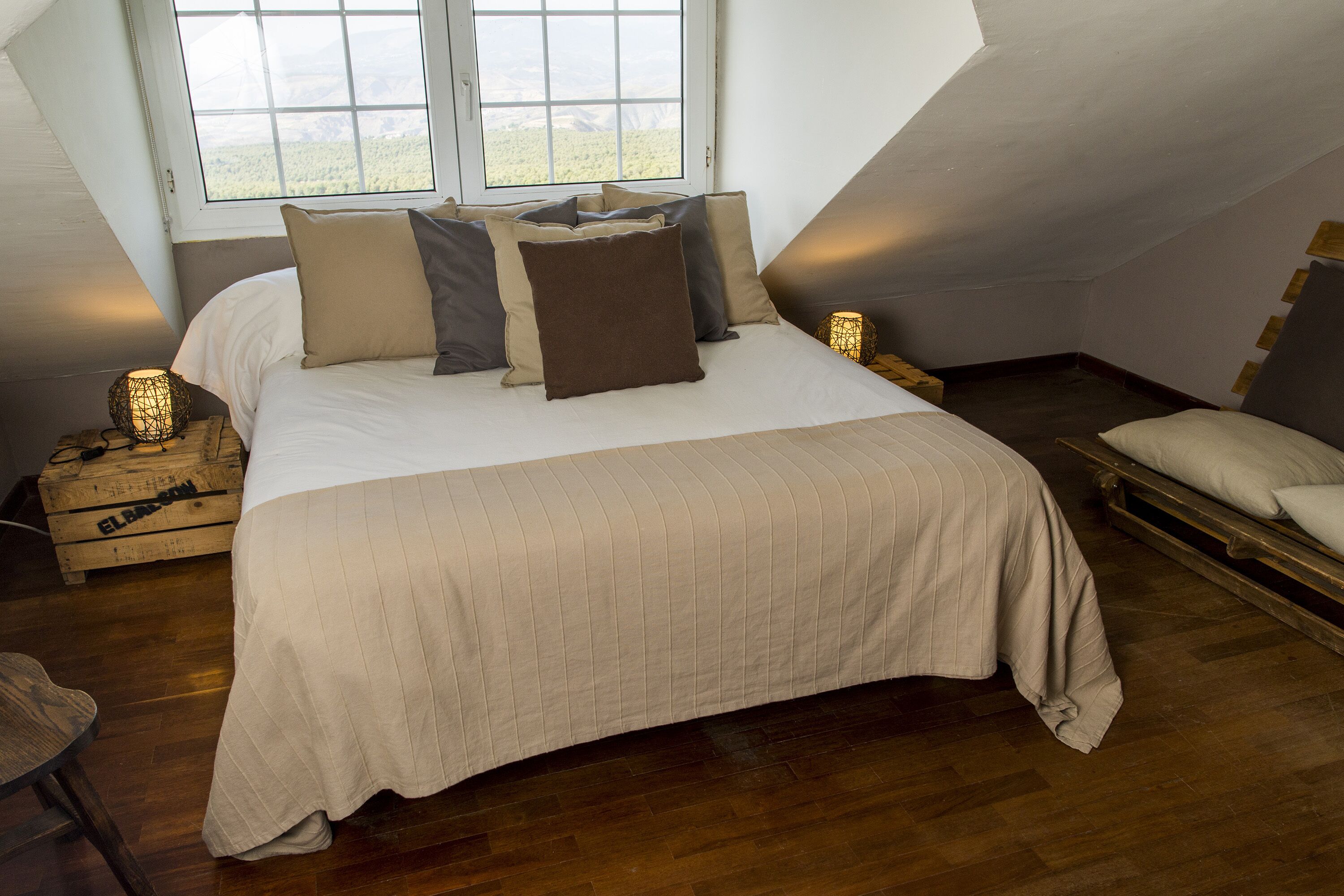 A bedroom with a large bed, wooden flooring, a window with a view, and a bedside table with a lamp.