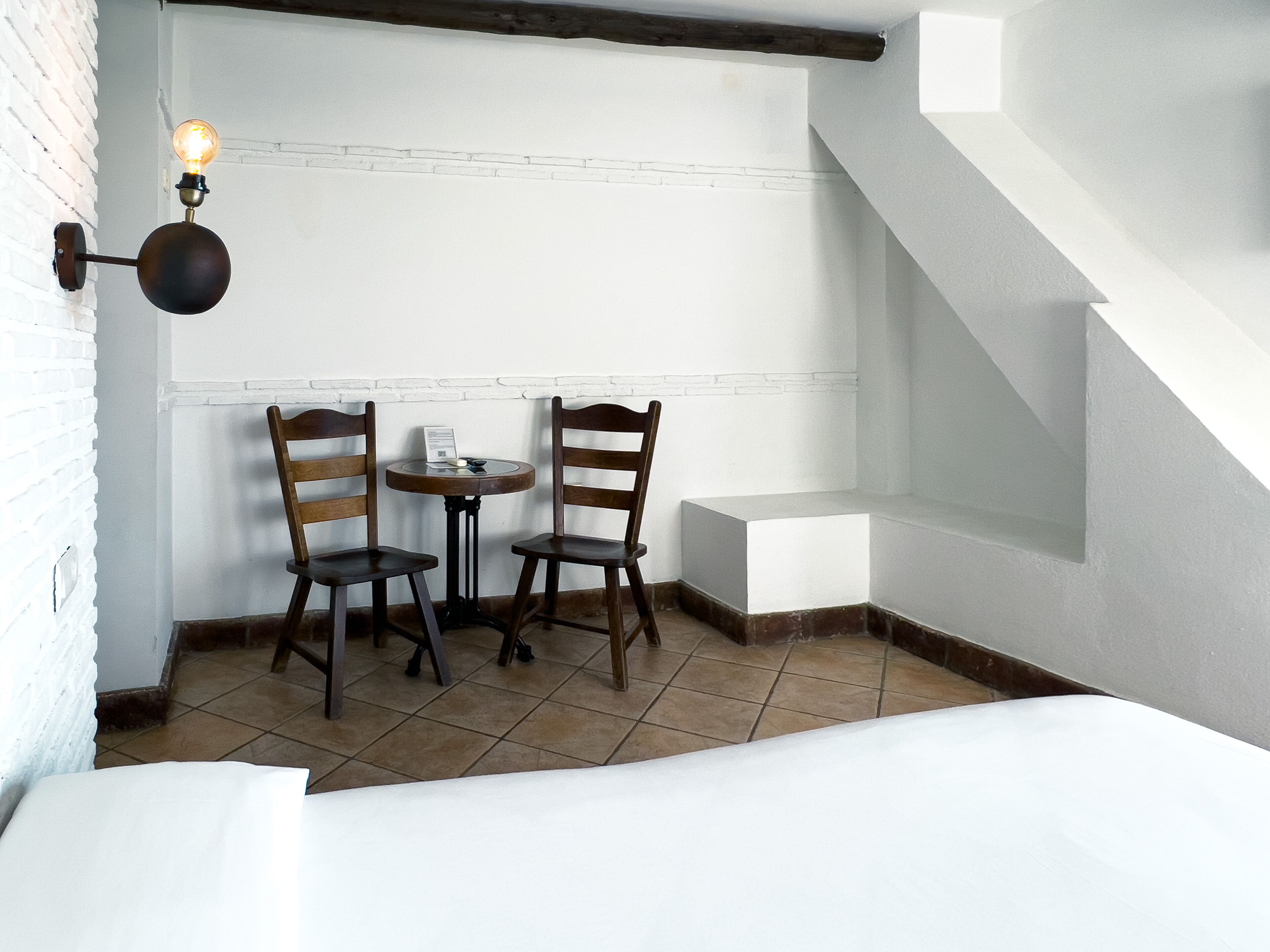 Una habitación pequeña y acogedora con dos sillas de madera, una mesita y una lámpara fijada a la pared.