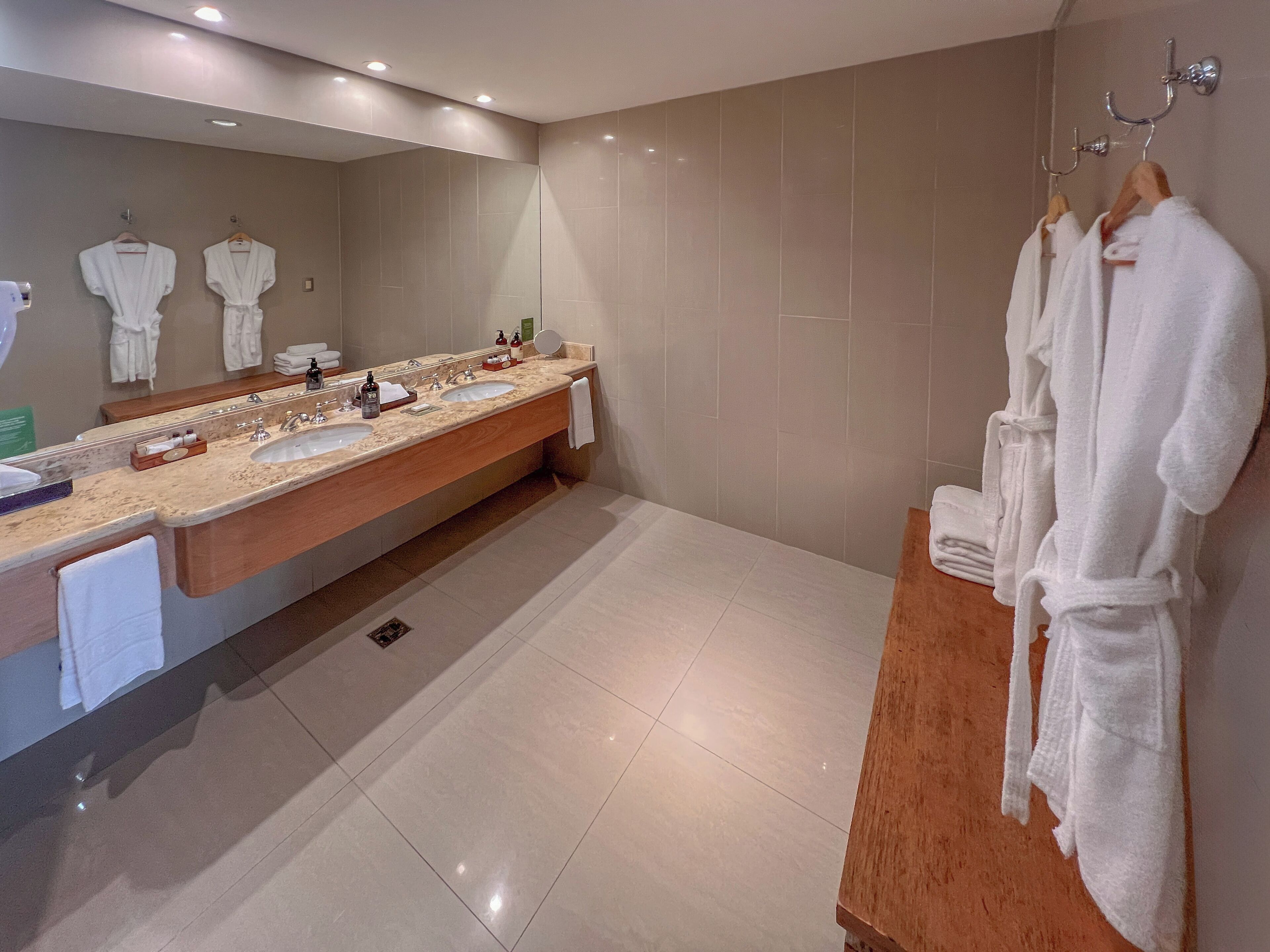 Un baño moderno con un amplio lavabo, dos lavamanos, un espejo y varias toallas y batas blancas.