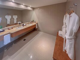 Un baño moderno con un amplio lavabo, dos lavamanos, un espejo y varias toallas y batas blancas.