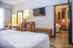 A hotel room with two beds, a wooden desk, a chair, a TV, and a bathroom visible through an open door.