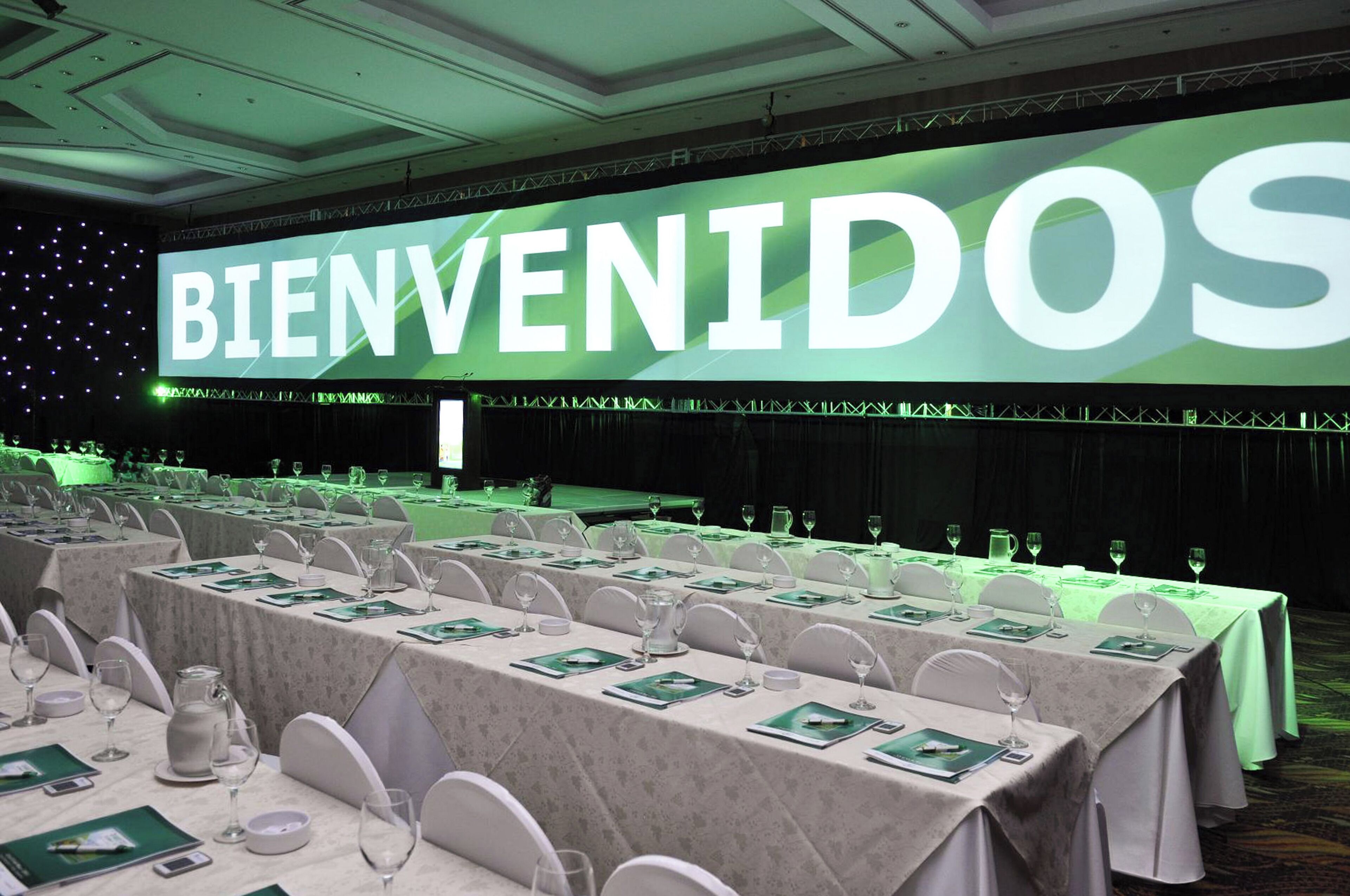 A conference room with tables set for a formal event, a large screen displaying "BIENVENIDOS" in the background, and a stage with a curtain.