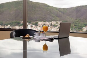 Una mesa con una computadora portátil, un vaso de jugo de naranja, una taza y un plato con comida, con vistas a una ciudad y colinas al fondo.