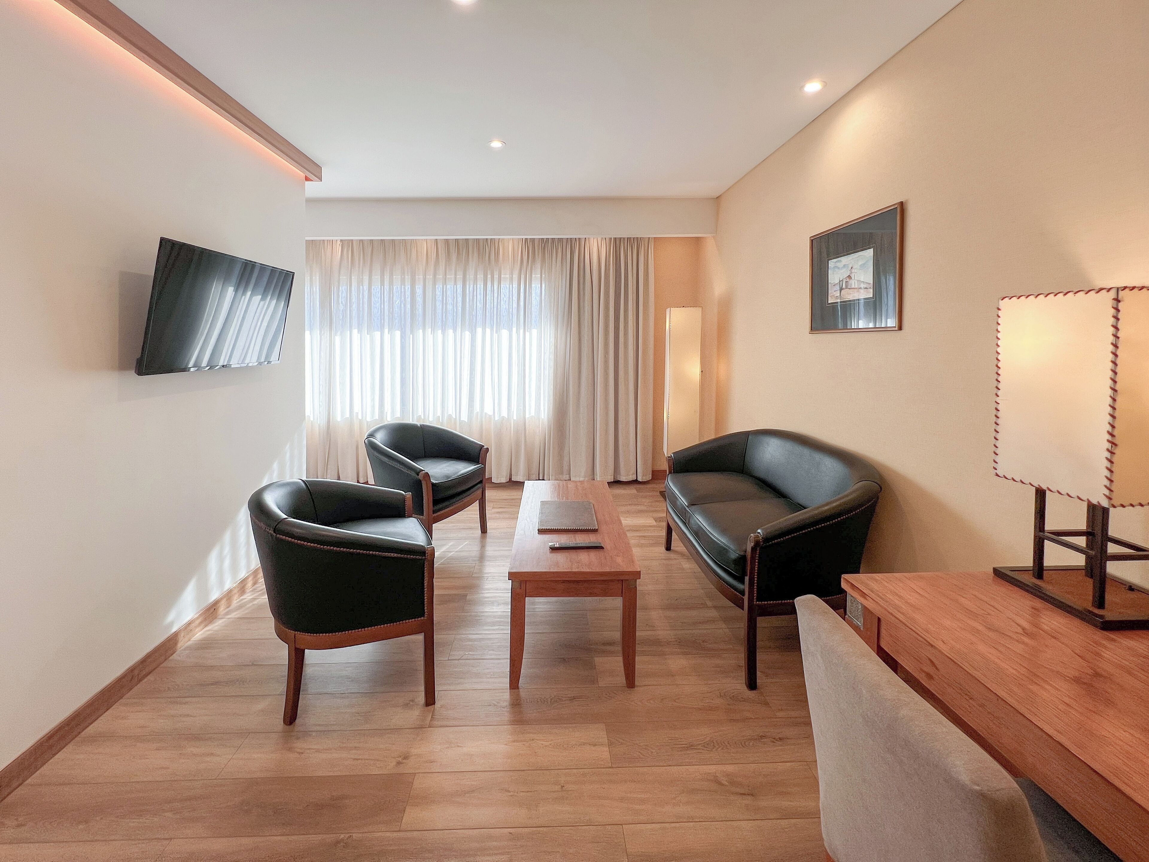 Une chambre d’hôtel moderne avec un plancher en bois, deux canapés en cuir, une table basse en bois, un téléviseur fixé au mur et une grande fenêtre avec des rideaux.