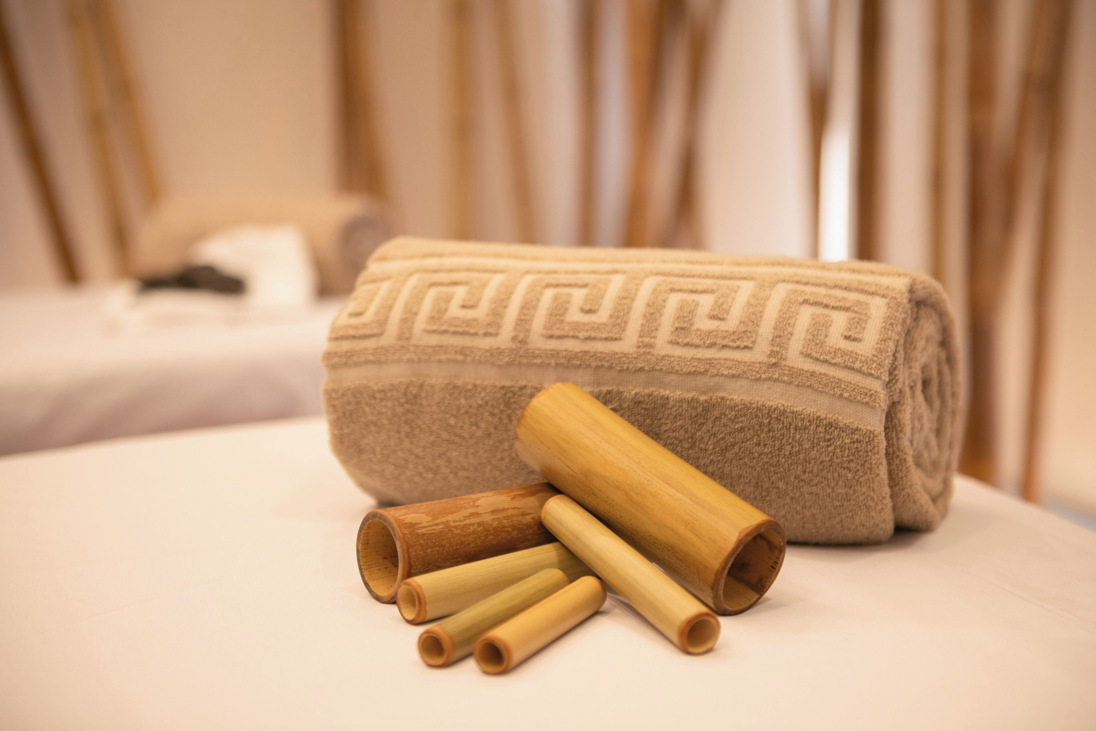 Une serviette beige pliée avec soin, ornée d’un motif géométrique, est posée sur un lit, accompagnée de plusieurs bâtonnets de bois cylindriques.