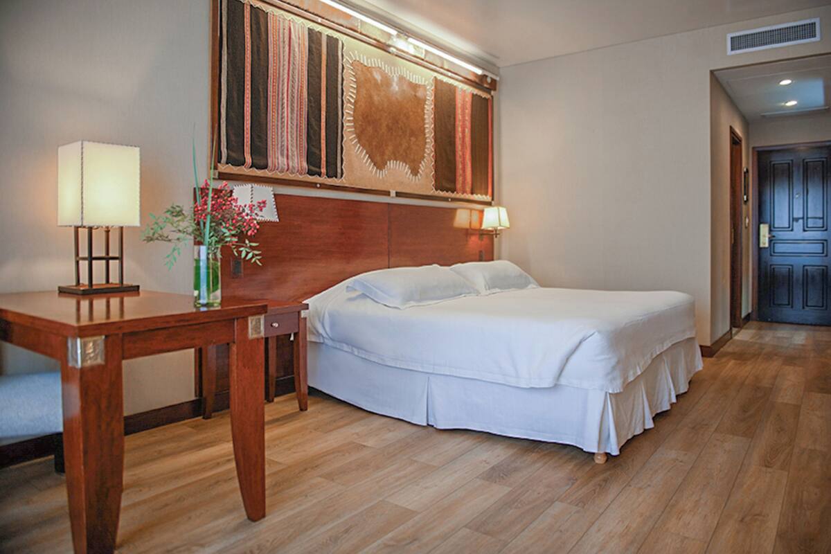 Una habitació d'hotel amb sòl de fusta, un llit amb roba de llit blanca, una tauleta de nit amb una làmpada, i una obra d'art penjada a la paret sobre el llit.