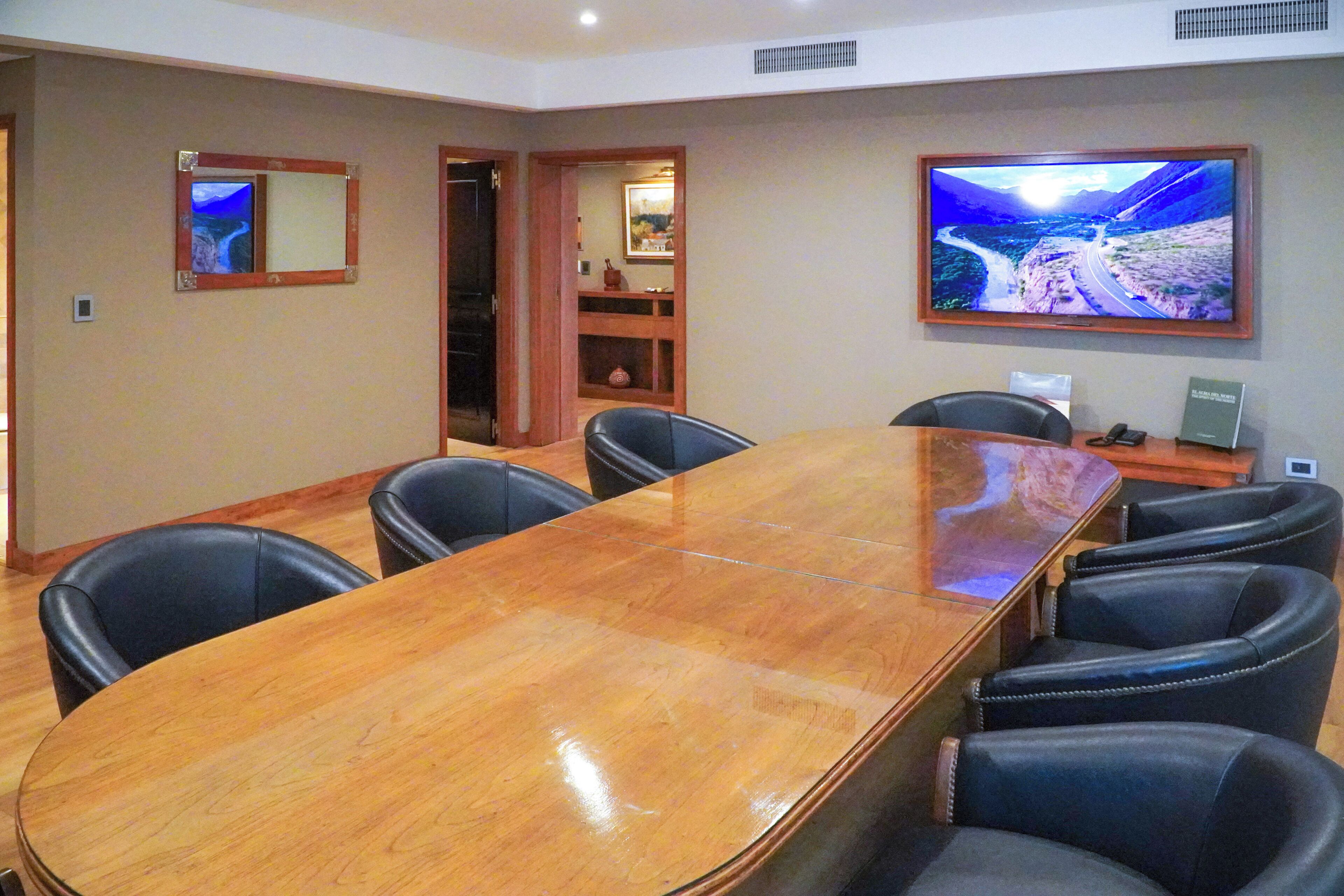 Una sala de conferencias con una larga mesa de madera, sillas de cuero negro, un televisor en la pared y un estante con adornos.