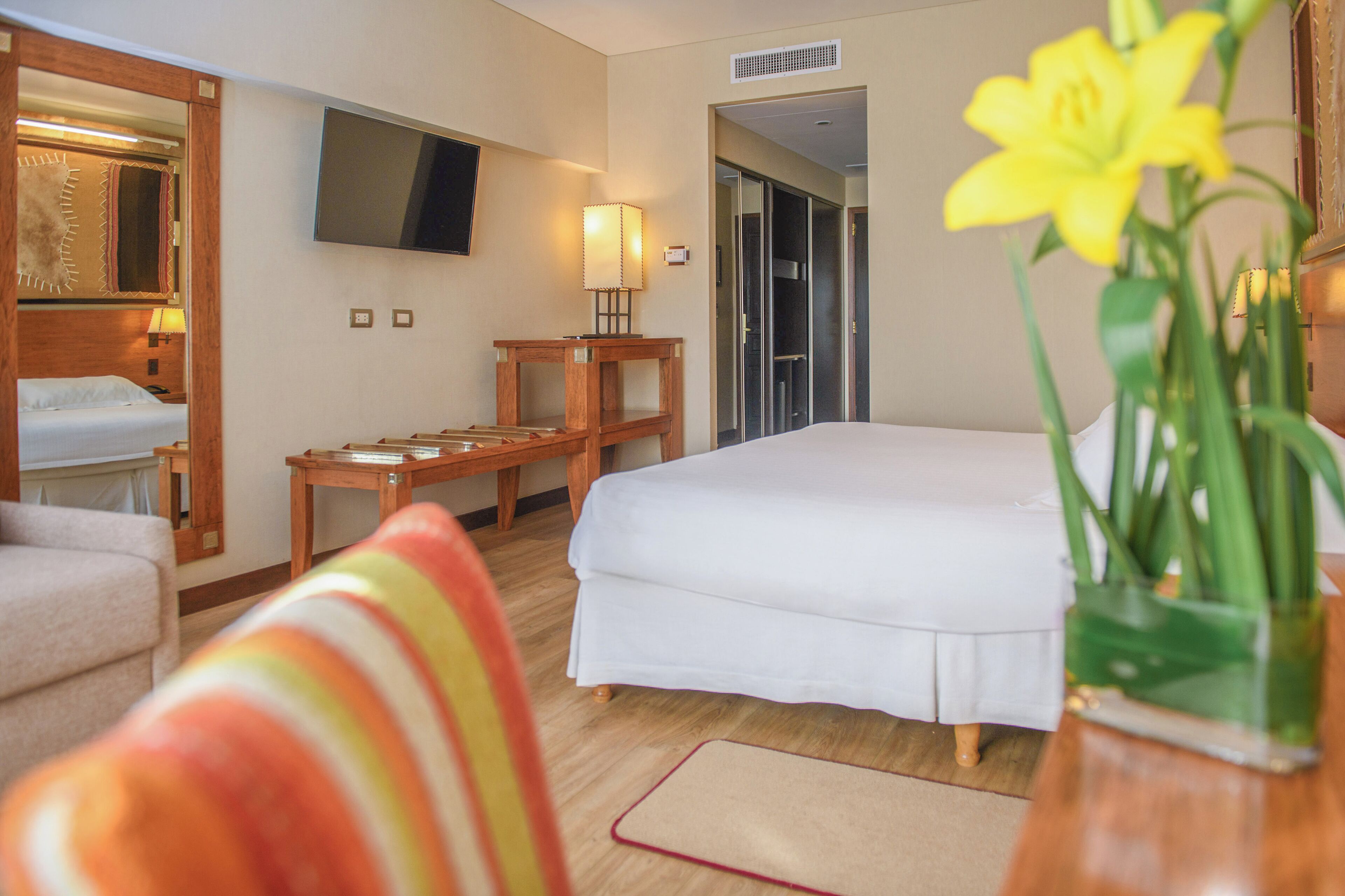 A hotel room with a bed, a TV mounted on the wall, a wooden nightstand, a lamp, and a vase with flowers.