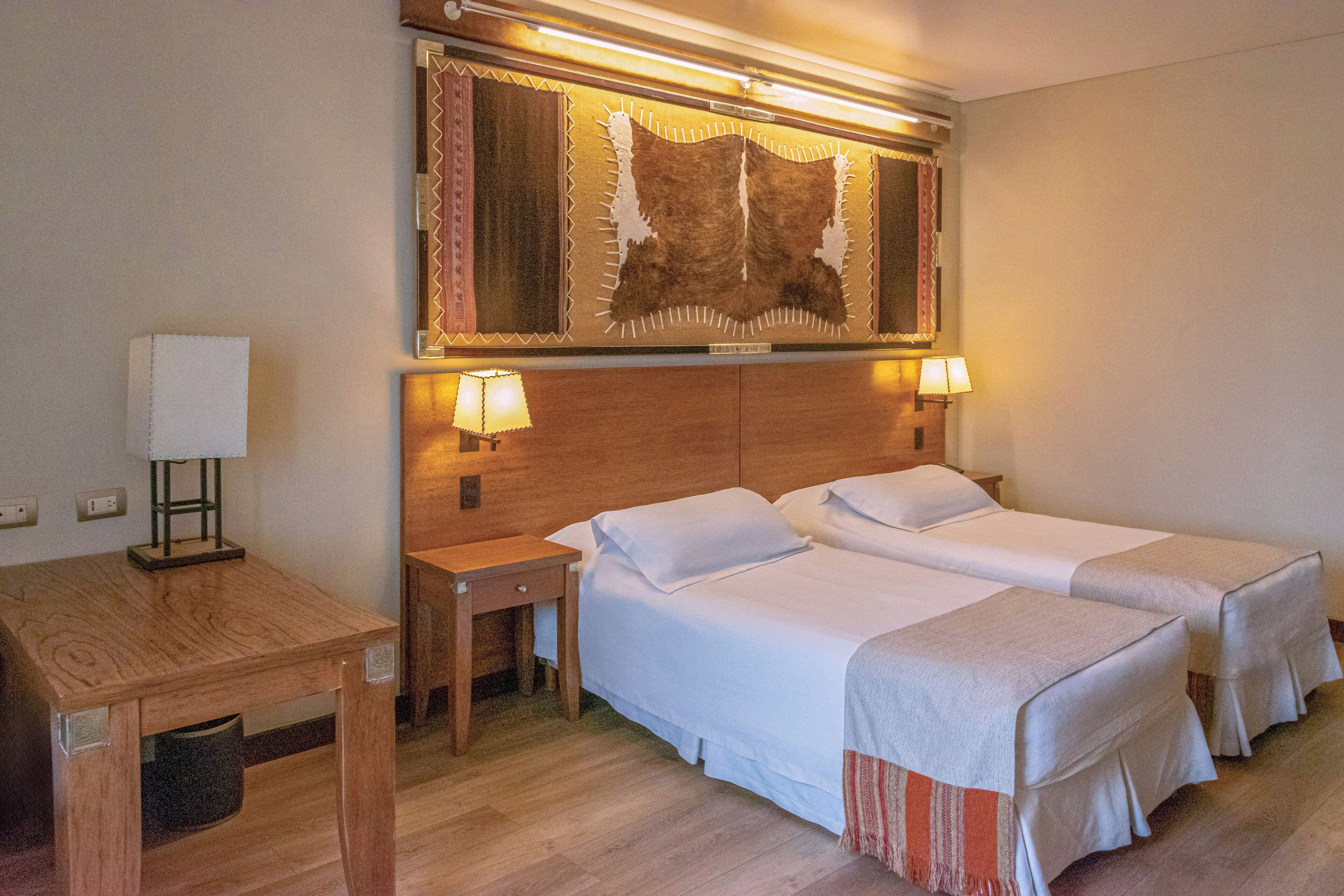 Ett hotellrum med två sängar, ett träbord vid sängen, ett skrivbord och en spegel som hänger på väggen.