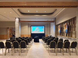 Una sala conferenze con file di sedie rivolte verso un grande schermo, pannelli di legno sulle pareti e pavimento piastrellato.
