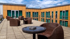 A rooftop terrace with wicker furniture, a round table, and a view of a building with green curtains. - Art Hotel William (Bratislava)