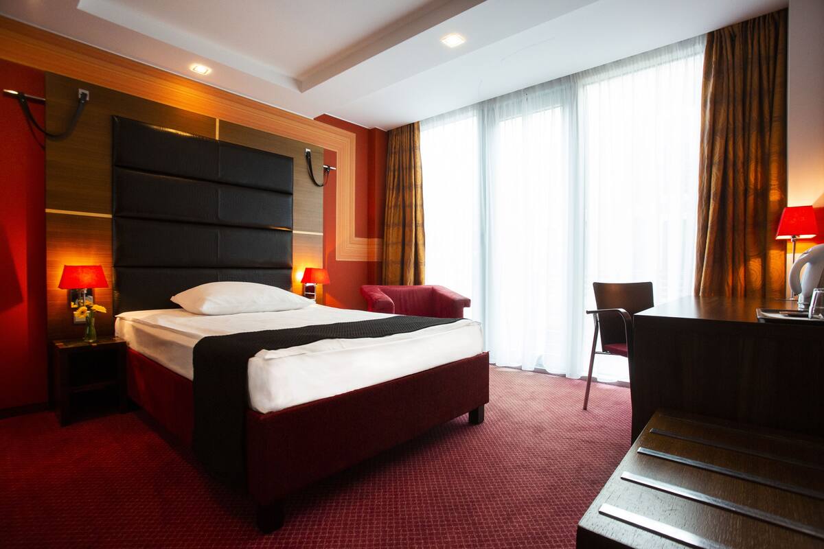 A hotel room with a large bed, a desk, and a chair. There is a window with curtains, a lamp on the desk, and a red carpet.