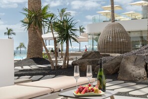 A beachside lounge with a table set with a bottle of champagne, glasses, and a fruit platter.